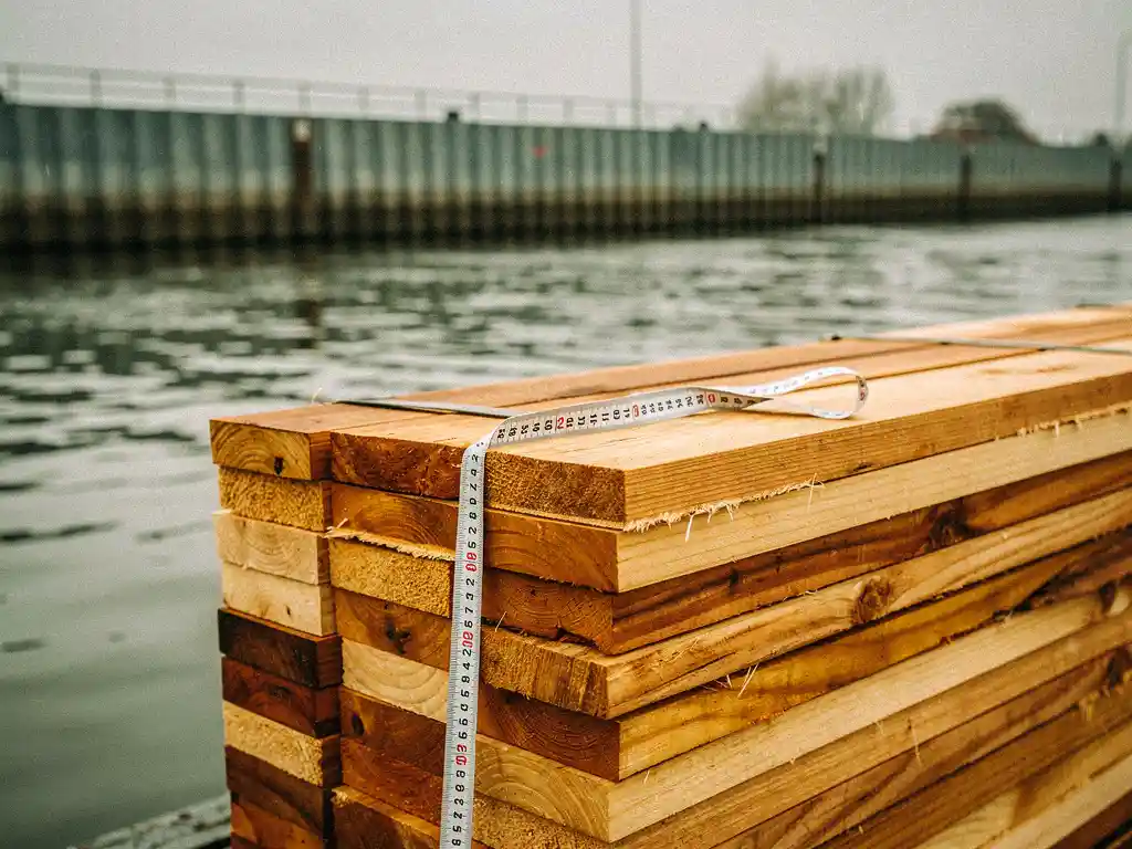 Ruw gezaagde houten planken met meetlint langs een Nederlandse waterweg, damwand zichtbaar langs de oever.